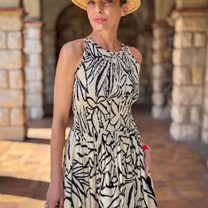 Può includere: Una donna indossa un cappello di paglia con una fascia blu navy e un vestito senza maniche color crema con un motivo a foglie nere. L'abito ha un corpetto aderente e una gonna fluida. Tiene in mano una borsa intrecciata.