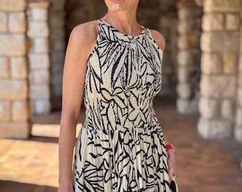 May include: A woman wearing a straw hat with a navy band and a sleeveless, cream-colored dress with a black leaf pattern. The dress has a fitted bodice and a flowing skirt. She is holding a woven handbag.