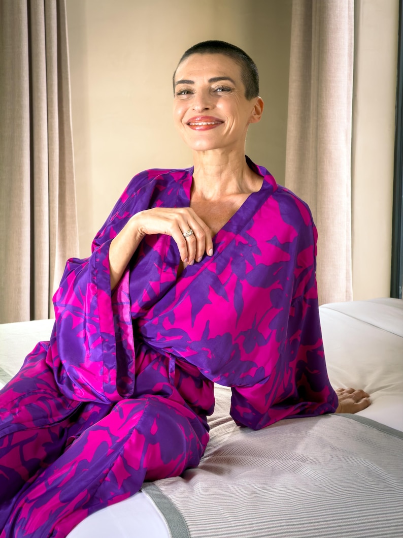 May include: A vibrant purple and fuchsia floral print kimono-style robe. The robe features a wide collar and long sleeves. The person is sitting on a bed with a striped comforter. The robe's bold colors create a striking visual.