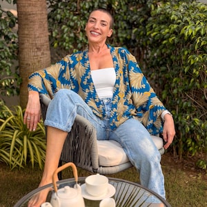May include: A blue and gold patterned kimono-style jacket. The jacket is worn over a white top and light blue jeans. A white teapot, teacup, and creamer sit on a glass table. The scene is set outdoors with greenery.