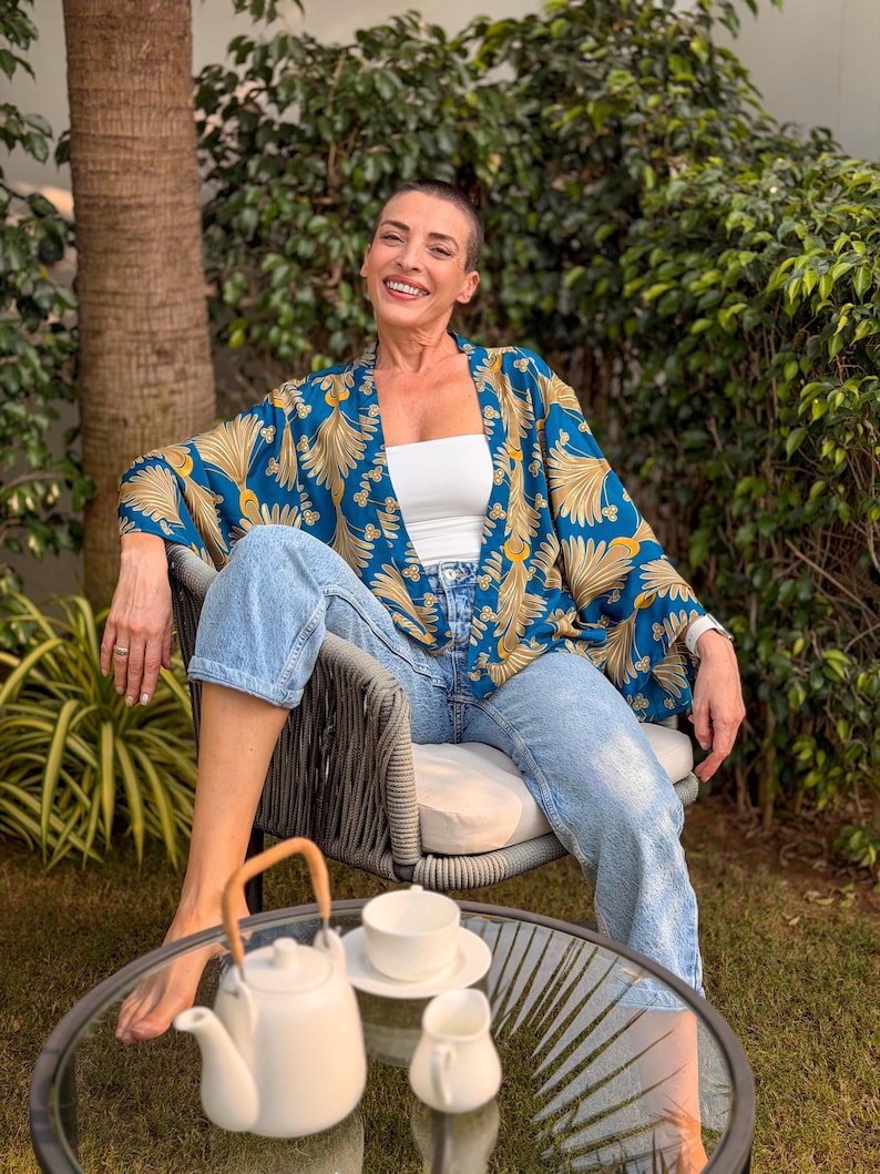 May include: A blue and gold patterned kimono-style jacket. The jacket is worn over a white top and light blue jeans. A white teapot, teacup, and creamer sit on a glass table. The scene is set outdoors with greenery.