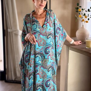 May include: A flowing, turquoise paisley print caftan dress with a deep V-neck. The dress features a vibrant pattern of red, blue, and black paisley designs. The sleeves are wide and the dress appears to be made of a lightweight fabric, suitable for warm weather.