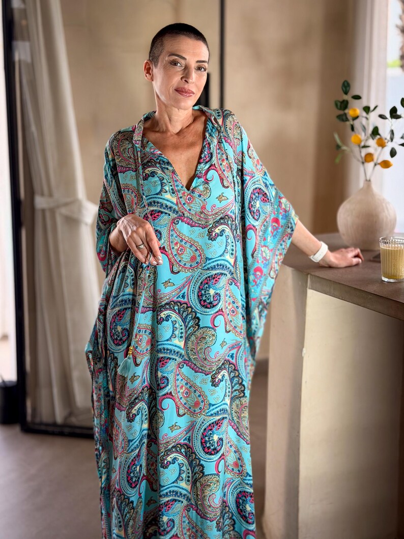 May include: A flowing, turquoise paisley print caftan dress with a deep V-neck. The dress features a vibrant pattern of red, blue, and black paisley designs. The sleeves are wide and the dress appears to be made of a lightweight fabric, suitable for warm weather.
