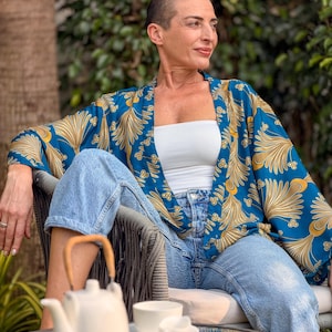 May include: A teal kimono with a gold floral pattern. The kimono is worn over a white tube top and light blue jeans. A white teapot, cup, and creamer sit on a glass table. The person is sitting in a gray chair.