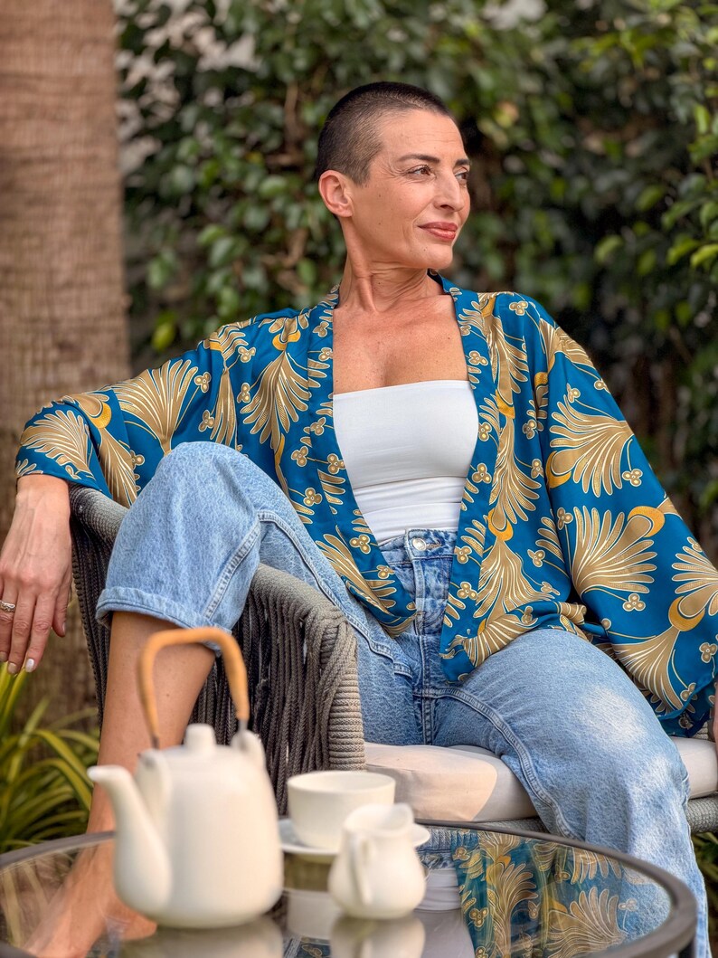 May include: A teal kimono with a gold floral pattern. The kimono is worn over a white tube top and light blue jeans. A white teapot, cup, and creamer sit on a glass table. The person is sitting in a gray chair.