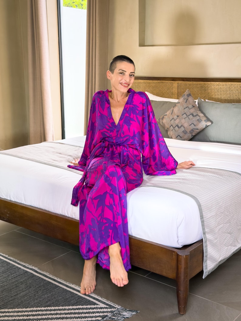 May include: A vibrant, long-sleeved robe in a pink and purple floral pattern. The robe is tied at the waist and has wide sleeves. The person is sitting on a bed with a wooden frame.