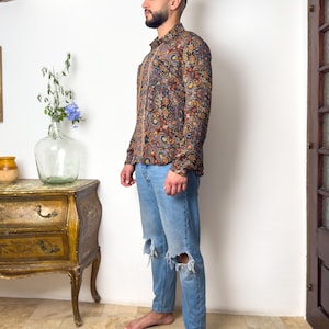 May include: A man wearing a long-sleeved shirt with a paisley pattern in shades of brown, blue, and orange. He is also wearing blue jeans with distressed knees.