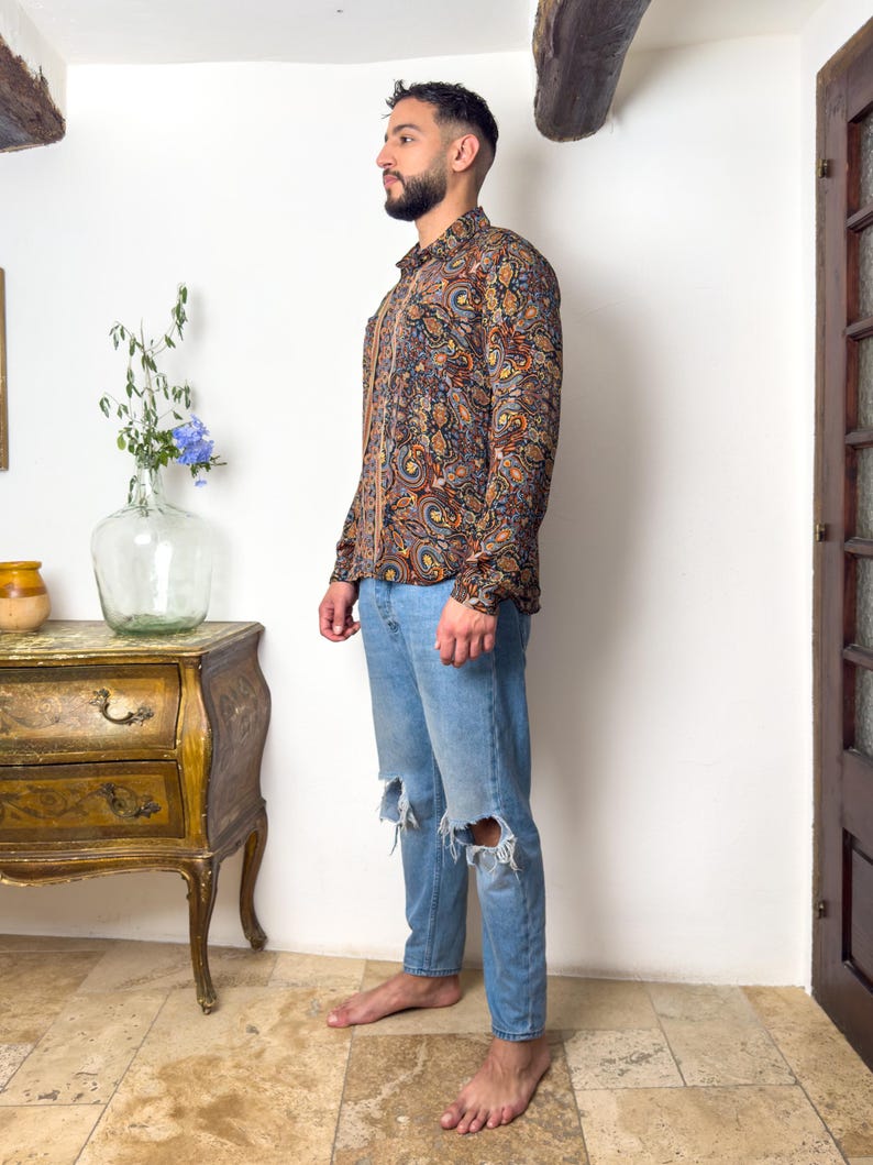 May include: A man wearing a long-sleeved shirt with a paisley pattern in shades of brown, blue, and orange. He is also wearing blue jeans with distressed knees.