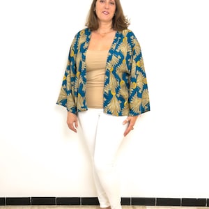 May include: A woman wearing a teal kimono with a gold leaf pattern, a beige top, and white pants. The kimono has wide sleeves. She is also wearing silver sandals. The background is a plain white wall.