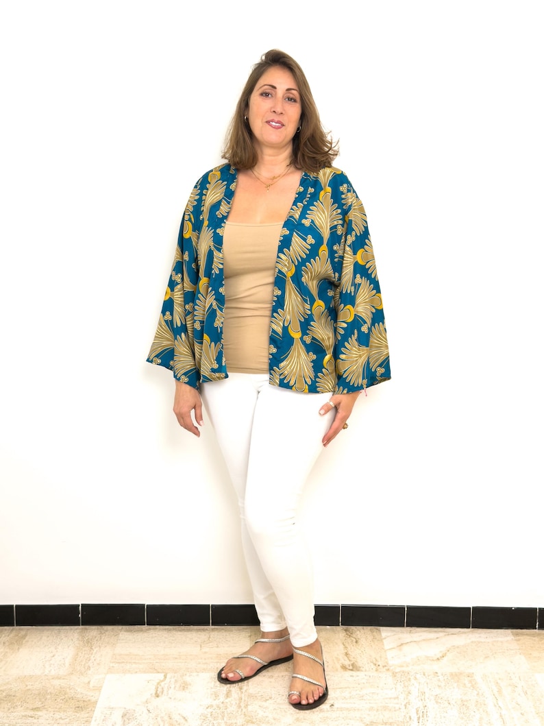 May include: A woman wearing a teal kimono with a gold leaf pattern, a beige top, and white pants. The kimono has wide sleeves. She is also wearing silver sandals. The background is a plain white wall.