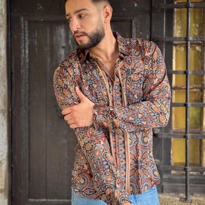 May include: A man wearing a long-sleeved shirt with a paisley pattern in shades of brown, blue, and orange. The shirt has a button-down collar and a relaxed fit. He is also wearing blue jeans.