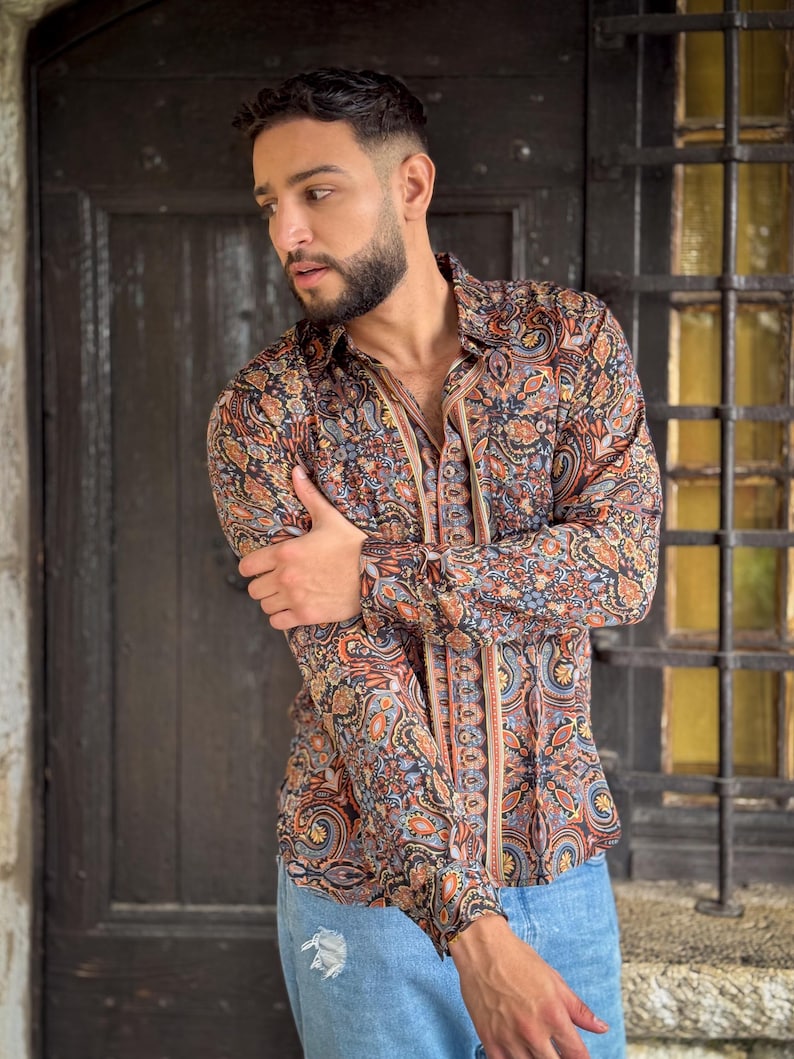 May include: A man wearing a long-sleeved shirt with a paisley pattern in shades of brown, blue, and orange. The shirt has a button-down collar and a relaxed fit. He is also wearing blue jeans.