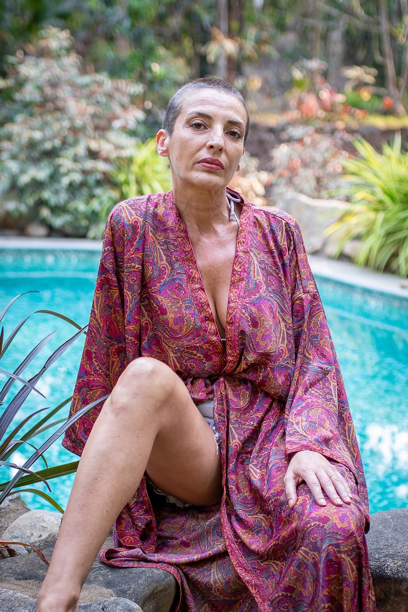 Peut inclure: Une femme portant une longue robe de chambre &agrave; fleurs rose et or. Elle est assise au bord d'une piscine, les jambes crois&eacute;es et les mains pos&eacute;es sur ses genoux.