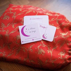 May include: A red drawstring bag with a gold floral pattern, accompanied by three small white cards. The cards feature text and a crescent moon design. One card reads "Un Grand Merci!", another "L'Atelier de Sonja", and the third says "Thanks!"