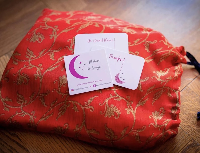 May include: A red drawstring bag with a gold floral pattern, accompanied by three small white cards. The cards feature text and a crescent moon design. One card reads "Un Grand Merci!", another "L'Atelier de Sonja", and the third says "Thanks!"