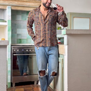 May include: A man wearing a brown and beige patterned long-sleeve shirt with a light blue denim shirt with distressed knees. He is standing in a kitchen and holding a glass of red wine.