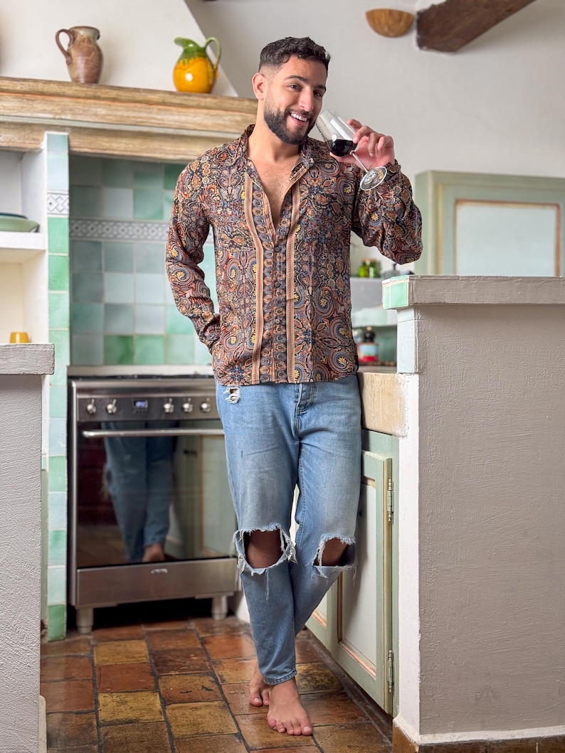 May include: A man wearing a brown and beige patterned long-sleeve shirt with a light blue denim shirt with distressed knees. He is standing in a kitchen and holding a glass of red wine.