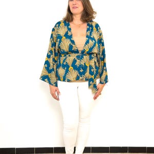 May include: A woman wearing a teal and gold patterned kimono-style top with a matching belt. She is also wearing a beige top, white pants, and silver sandals. The background is a plain white wall.