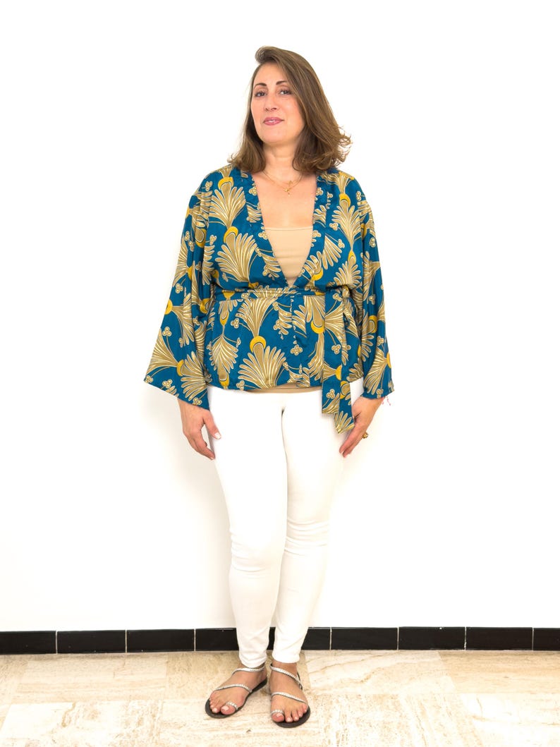 May include: A woman wearing a teal and gold patterned kimono-style top with a matching belt. She is also wearing a beige top, white pants, and silver sandals. The background is a plain white wall.