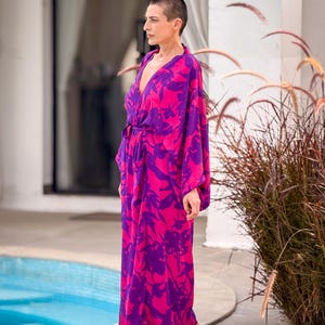 May include: A long, flowing robe in vibrant pink and purple, featuring a floral pattern. The robe has a V-neck and a tie at the waist. The sleeves are long and loose. The garment is worn outdoors near a pool.