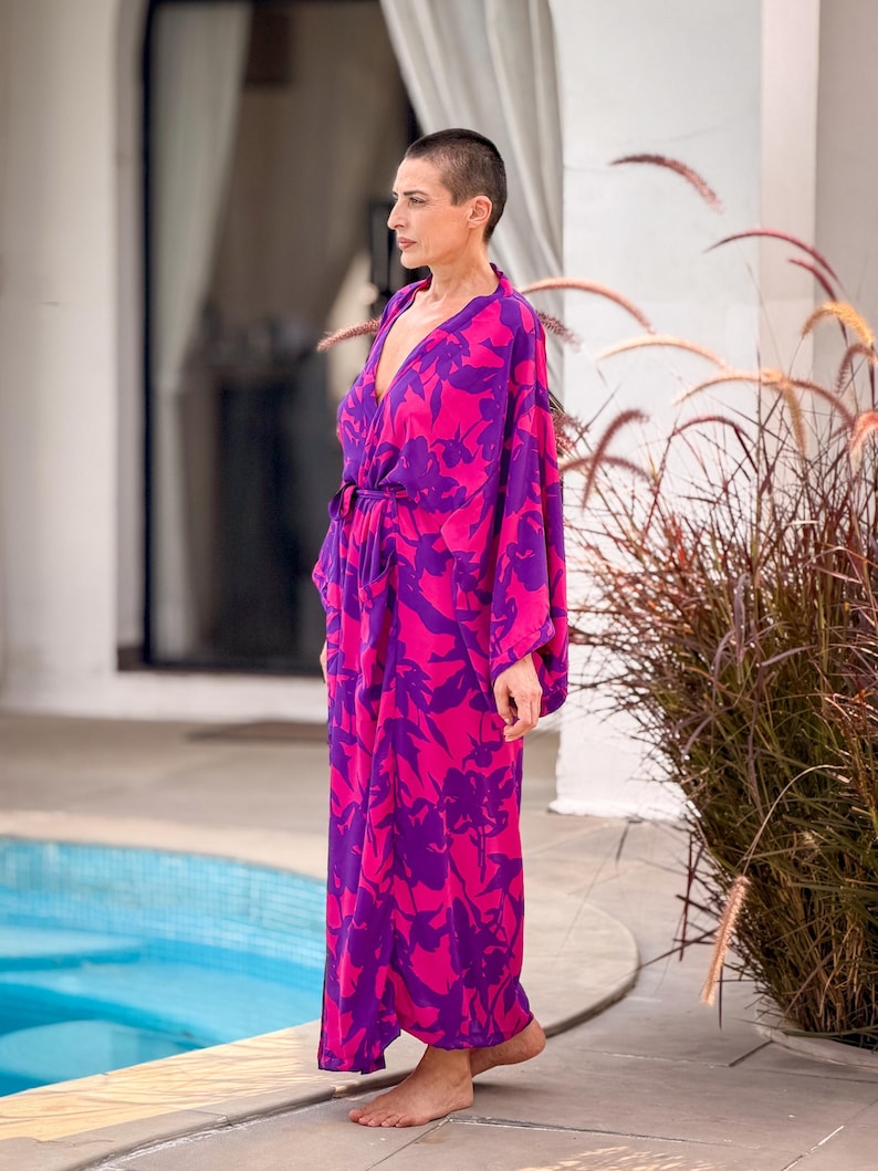 May include: A long, flowing robe in vibrant pink and purple, featuring a floral pattern. The robe has a V-neck and a tie at the waist. The sleeves are long and loose. The garment is worn outdoors near a pool.