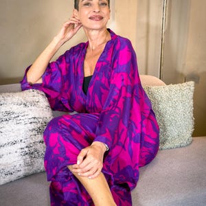 May include: A vibrant, flowing kimono in shades of fuchsia and purple, featuring a bold floral pattern. The garment is worn open, revealing a black top underneath. The model is seated on a gray sofa, creating a striking contrast with the colorful kimono.