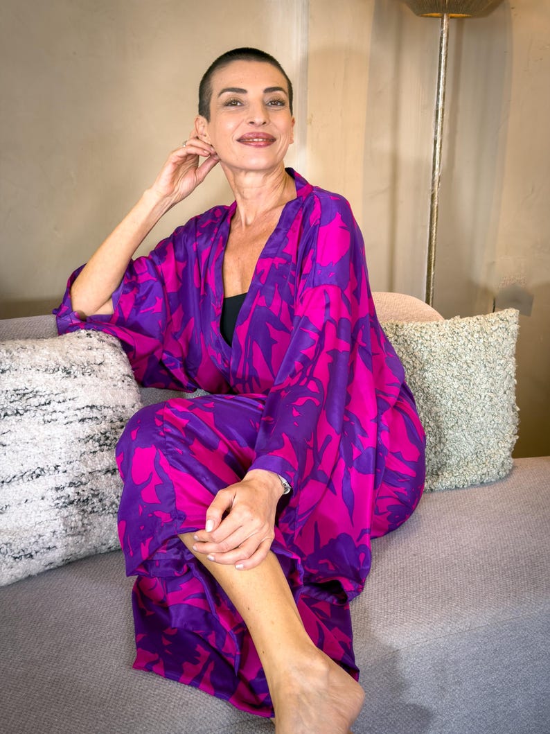 May include: A vibrant, flowing kimono in shades of fuchsia and purple, featuring a bold floral pattern. The garment is worn open, revealing a black top underneath. The model is seated on a gray sofa, creating a striking contrast with the colorful kimono.