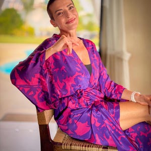 May include: A woman wearing a purple and pink floral patterned robe. The robe is tied at the waist and has wide sleeves. She is sitting on a wooden chair with woven seat and back. The background is blurred, with a view of greenery.