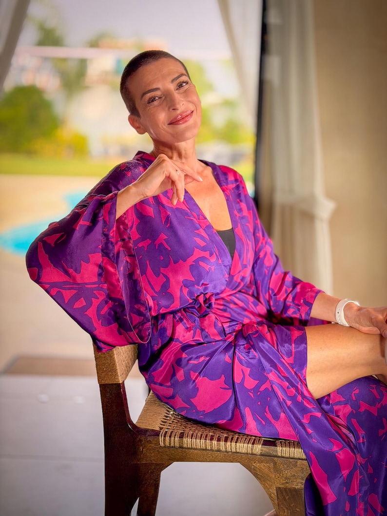 May include: A woman wearing a purple and pink floral patterned robe. The robe is tied at the waist and has wide sleeves. She is sitting on a wooden chair with woven seat and back. The background is blurred, with a view of greenery.