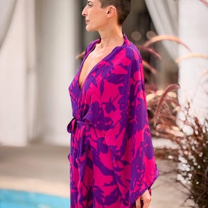 May include: A vibrant, flowing robe in shades of fuchsia and purple, adorned with a floral pattern. The robe features a wrap design with a matching tie belt and wide sleeves. The model is wearing the robe outdoors.