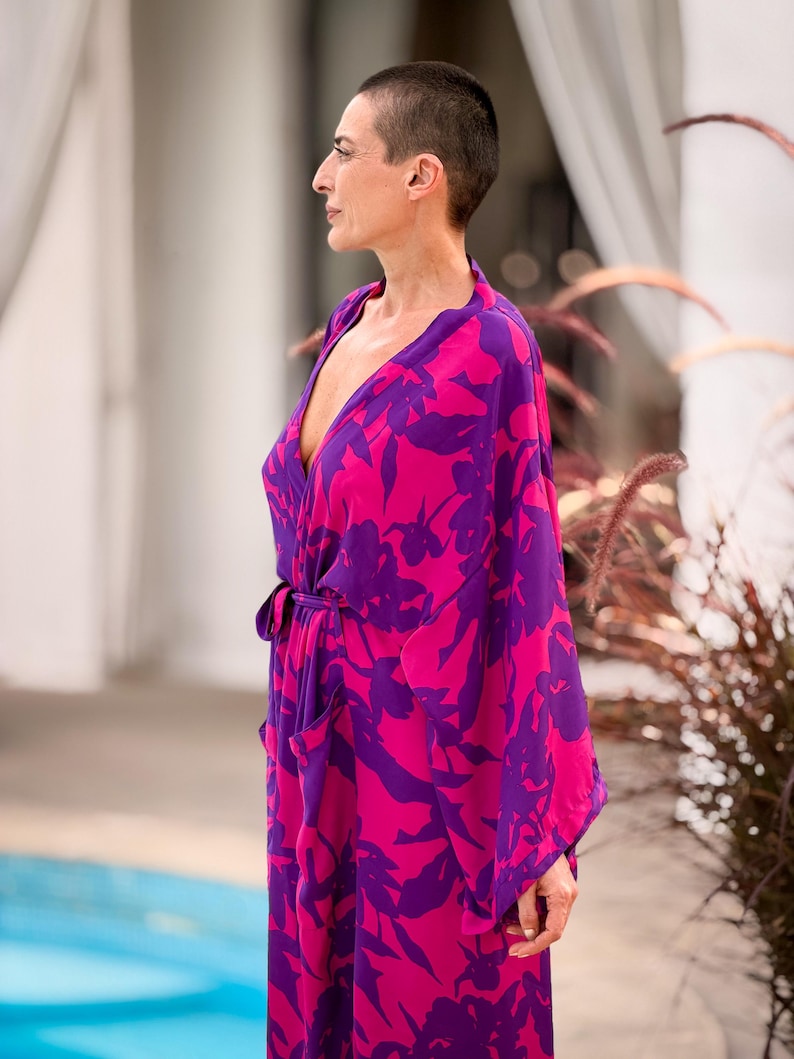 May include: A vibrant, flowing robe in shades of fuchsia and purple, adorned with a floral pattern. The robe features a wrap design with a matching tie belt and wide sleeves. The model is wearing the robe outdoors.