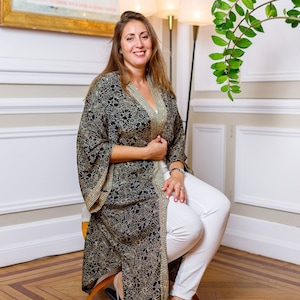 May include: A woman wearing a black and gold patterned kimono with white trousers and silver sandals. She is sitting on a stool in front of a white wall with a framed poster that reads "Cynthia Loemij & Mark Lorimer To Intimate".