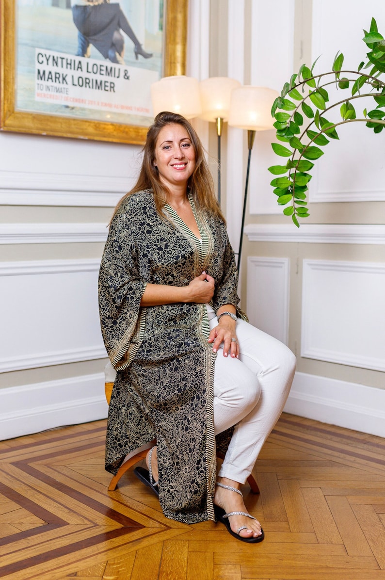 May include: A woman wearing a black and gold patterned kimono with white trousers and silver sandals. She is sitting on a stool in front of a white wall with a framed poster that reads "Cynthia Loemij & Mark Lorimer To Intimate".