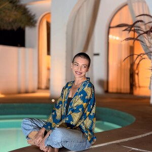 May include: A blue and gold patterned kimono-style jacket. The jacket is worn over a black top and paired with blue jeans. The person is sitting cross-legged near a pool, with a building in the background.