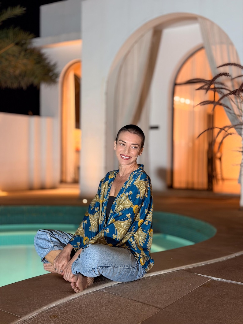 May include: A blue and gold patterned kimono-style jacket. The jacket is worn over a black top and paired with blue jeans. The person is sitting cross-legged near a pool, with a building in the background.