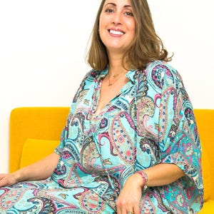 May include: A woman wearing a turquoise paisley print dress with pink and blue accents. The dress has a collared neckline and short sleeves. She is sitting on a yellow couch.