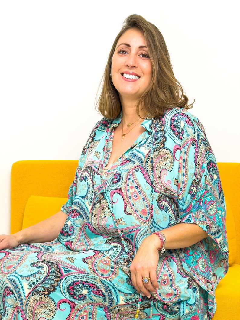 May include: A woman wearing a turquoise paisley print dress with pink and blue accents. The dress has a collared neckline and short sleeves. She is sitting on a yellow couch.