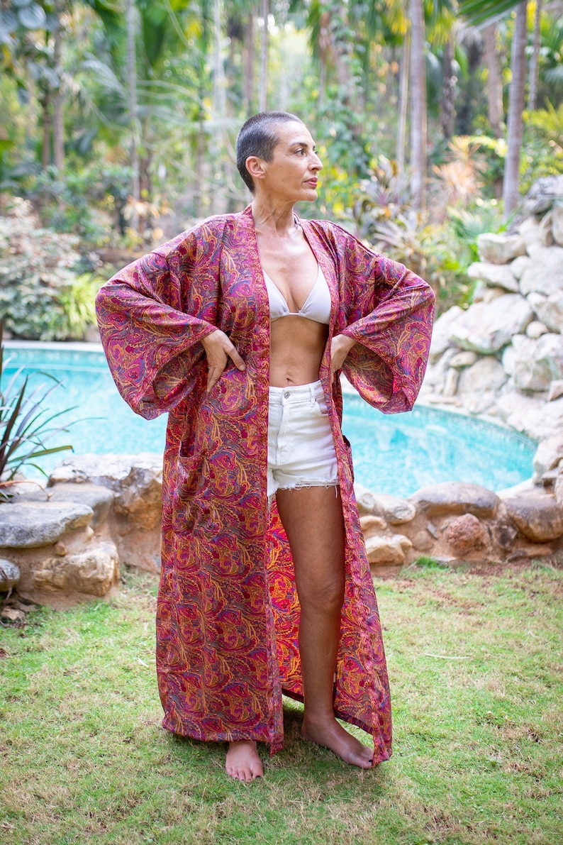 Peut inclure: Une femme portant un kimono &agrave; motifs rouge et or avec un haut de bikini blanc et un short en jean blanc. Elle se tient devant une piscine avec une v&eacute;g&eacute;tation luxuriante en arri&egrave;re-plan.