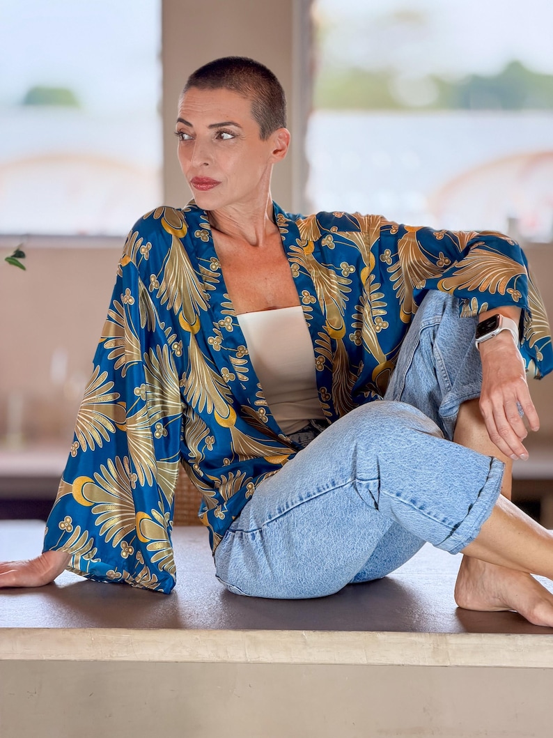 May include: A blue kimono-style robe with a gold floral pattern. The robe is worn over a white top and light blue jeans. The sleeves are long and flowing. The person is sitting on a dark surface.