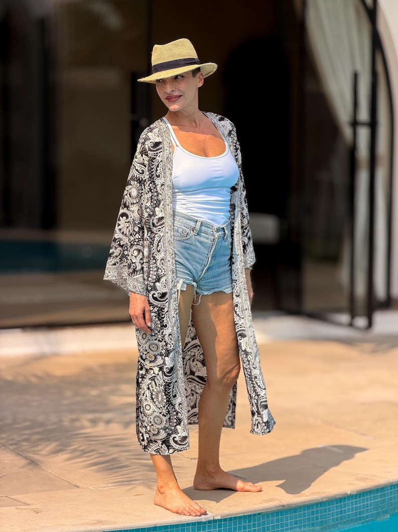 May include: A woman wearing a straw hat, white tank top, denim shorts, and a long black and white patterned kimono. The outfit is suitable for warm weather. The woman is standing near a swimming pool.