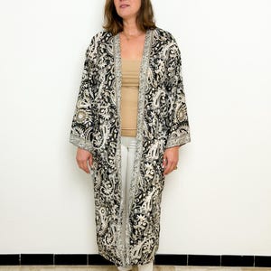 May include: A long, open-front kimono in black and cream paisley print. The kimono features wide sleeves and a silver trim along the edges. The model is wearing a beige top, white pants, and silver sandals.