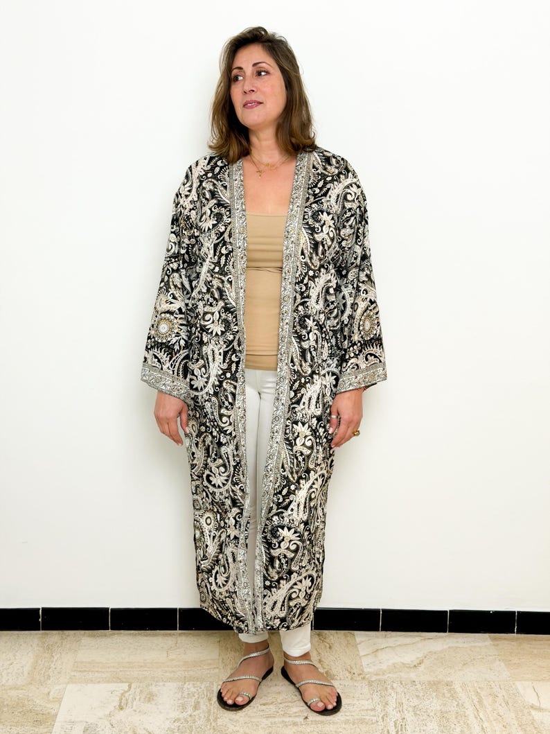 May include: A long, open-front kimono in black and cream paisley print. The kimono features wide sleeves and a silver trim along the edges. The model is wearing a beige top, white pants, and silver sandals.