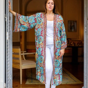 May include: A woman wearing a long, flowing, teal and red patterned kimono with white pants and silver sandals. The kimono has a gold trim and intricate floral designs.