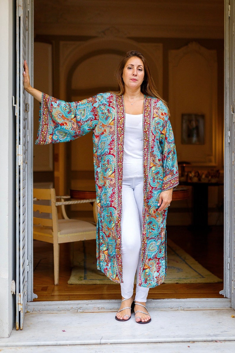 May include: A woman wearing a long, flowing, teal and red patterned kimono with white pants and silver sandals. The kimono has a gold trim and intricate floral designs.