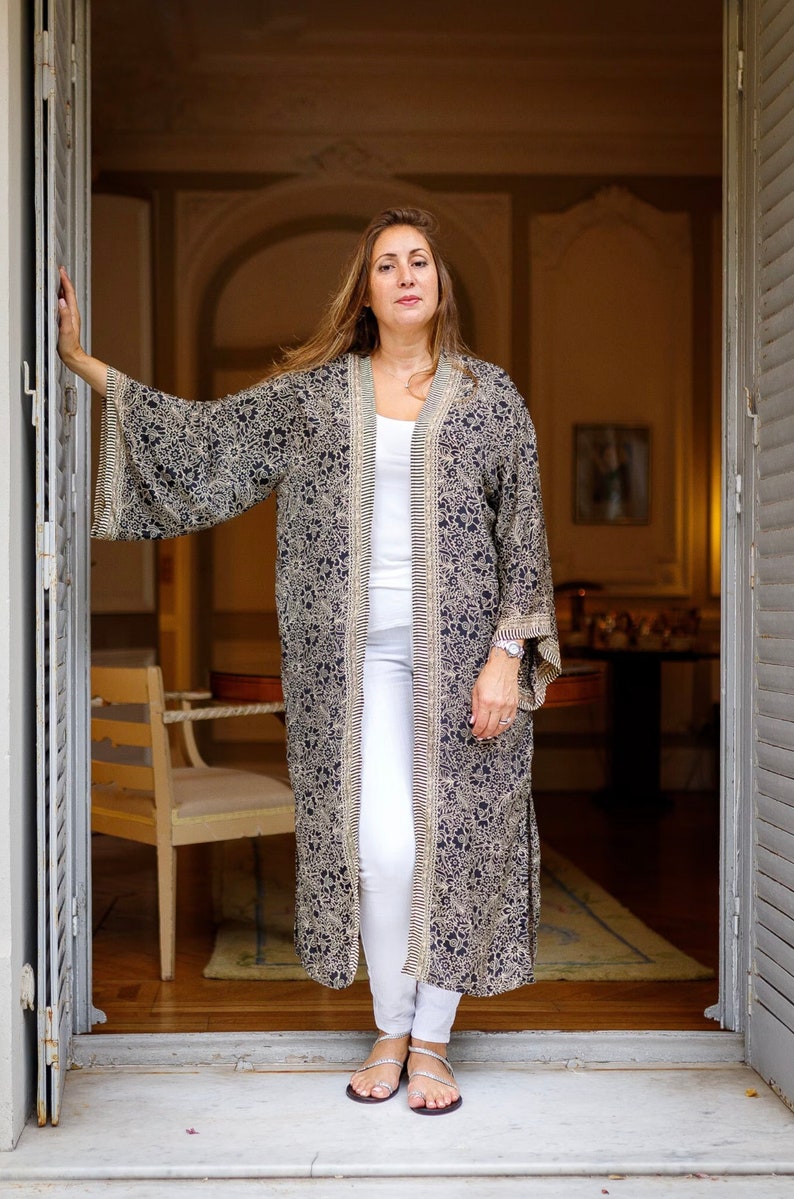 May include: A woman wearing a long, black and white patterned kimono with white trousers and silver sandals. She is standing in a doorway with a wooden floor and a white door frame.