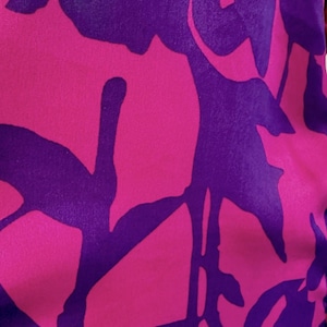 May include: Close-up of a fabric with a floral pattern. The design features large, stylized flowers in vibrant shades of pink and purple. The background is a deep purple, and the flowers are a bright pink, creating a striking contrast.