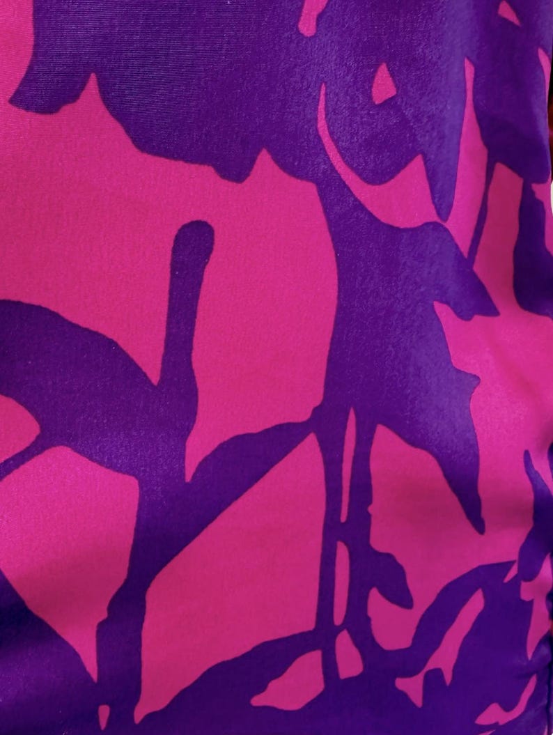 May include: Close-up of a fabric with a floral pattern. The design features large, stylized flowers in vibrant shades of pink and purple. The background is a deep purple, and the flowers are a bright pink, creating a striking contrast.