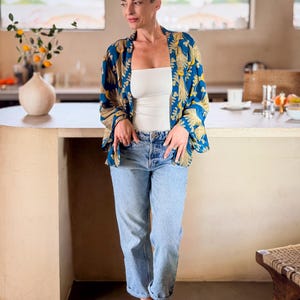 May include: A woman wearing a blue and gold patterned kimono over a white top and light blue jeans. The kimono features a botanical design. The woman is standing in a kitchen setting.