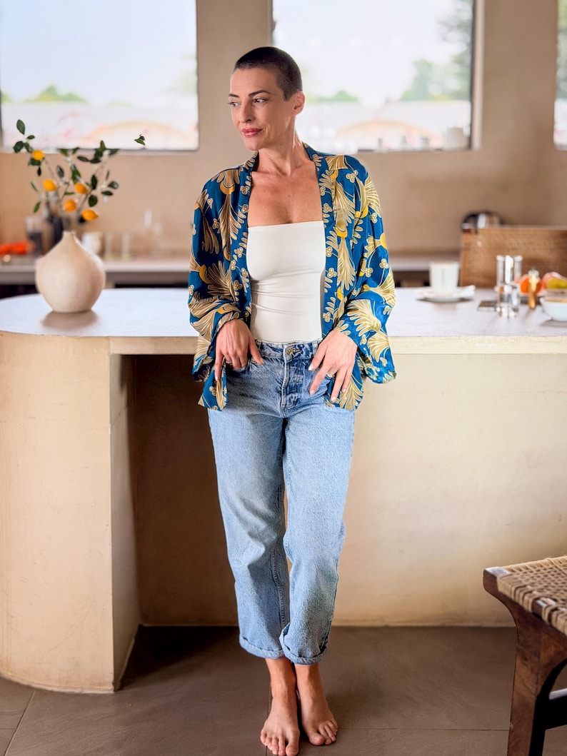 May include: A woman wearing a blue and gold patterned kimono over a white top and light blue jeans. The kimono features a botanical design. The woman is standing in a kitchen setting.
