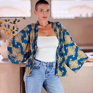 May include: A blue kimono-style jacket with a gold and white floral pattern. The jacket is worn over a white tube top and light blue jeans. The sleeves are wide and the fabric appears to be lightweight.
